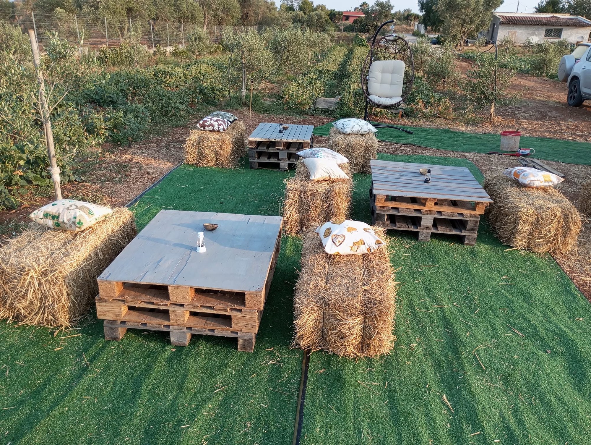 Tavoli di bancali e balle di fieno con cuscini su prato sintetico tra ulivi e orto di Agrifood da Francesco