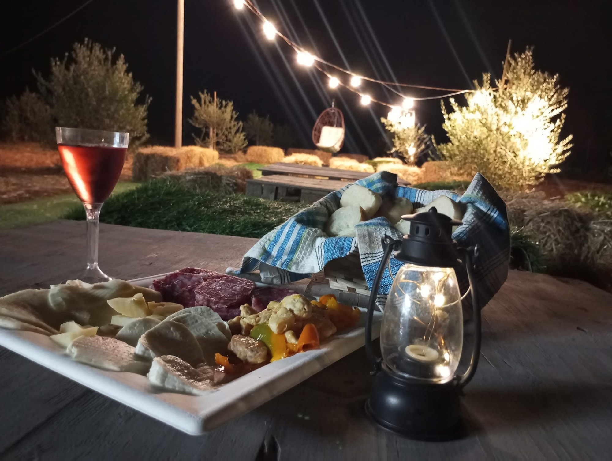 Piatto di salumi, formaggi e peperoni con vino rosato, pane e lanterna su tavolo in legno tra ulivi illuminati