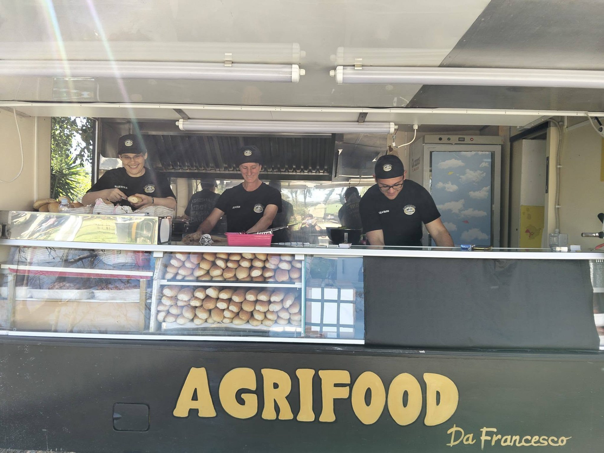 Tre addetti in maglietta e cappello neri preparano panini nel food truck con insegna gialla AGRIFOOD Da Francesco